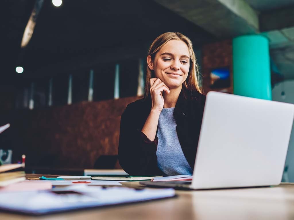 Žena sedí u stolu, soustředěná na svůj notebook, vedle má blok a pero