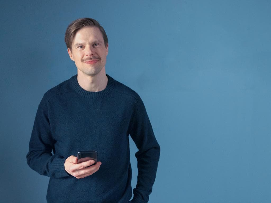 Matti v svetru drží mobilní telefon a dívá se na obrazovku