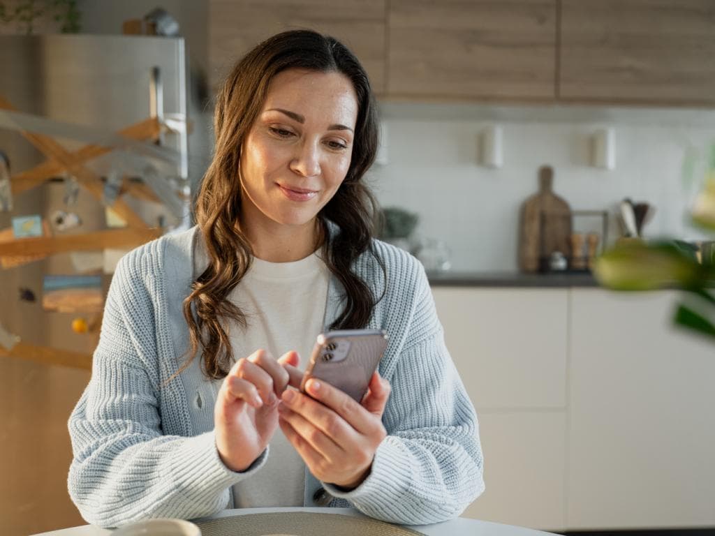 Žena sedí u stolu, soustředěná na svůj telefon
