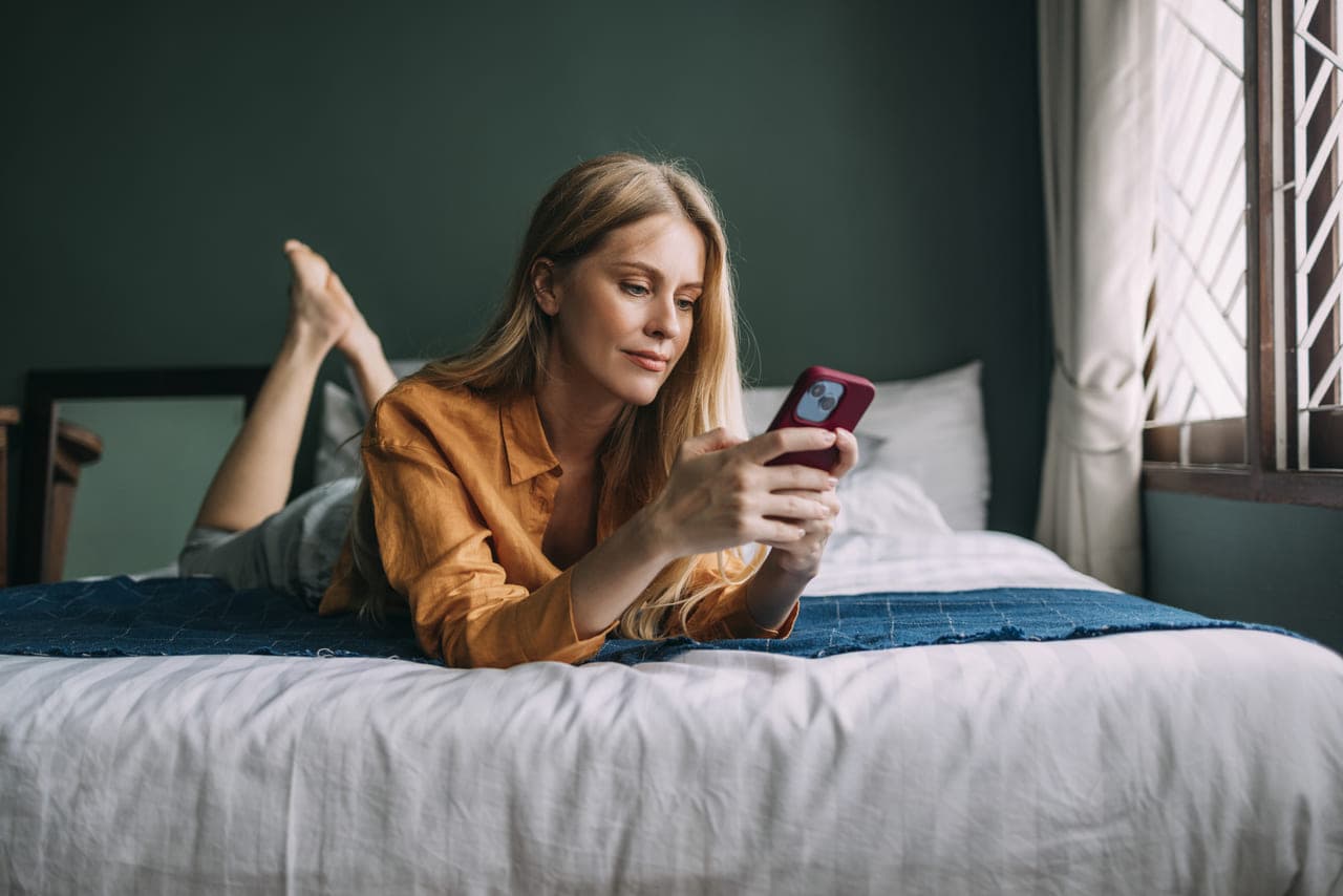 Žena leží na posteli, soustředěná na svůj telefon, obklopená měkkým povlečením a útulnou atmosférou.